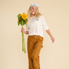 Load image into Gallery viewer, A woman wearing a Bluestone Lane tshirt and hat is smiling, holding a bunch of sunflowers
